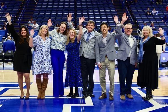 A group of eight people stands on a basketball court, smiling and waving at the camera. They are dressed in professional attire, with a mix of suits, dresses, and blazers. The background features rows of empty blue stadium seats, indicating a large indoor sports arena. The individuals appear to be celebrating or being recognized at an event.