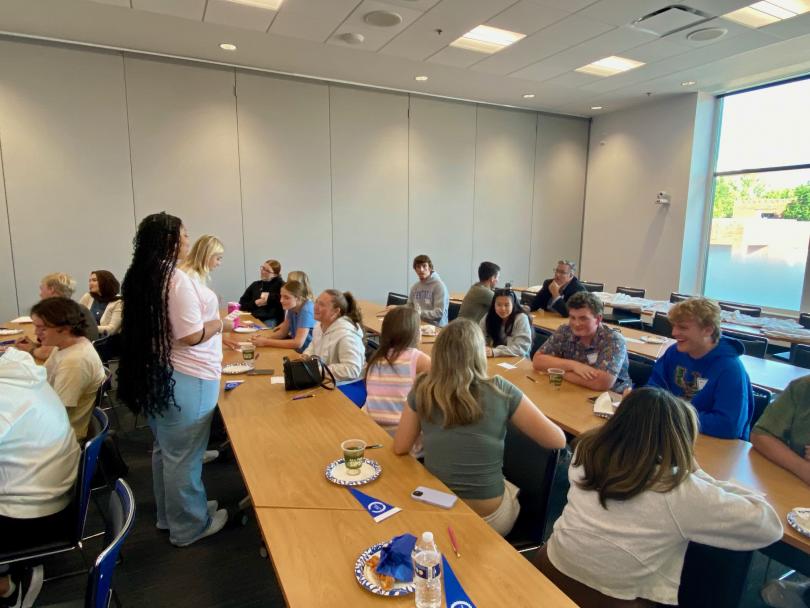 A gathering of students talk informally.  Most are casually seated and smiling.