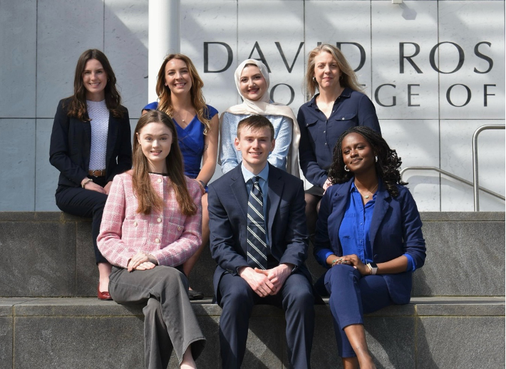 Seven professionally dressed people, four women standing and three seated, pose on outdoor steps in front of a building with partial signage reading "DAVID ROS COLLEGE OF.